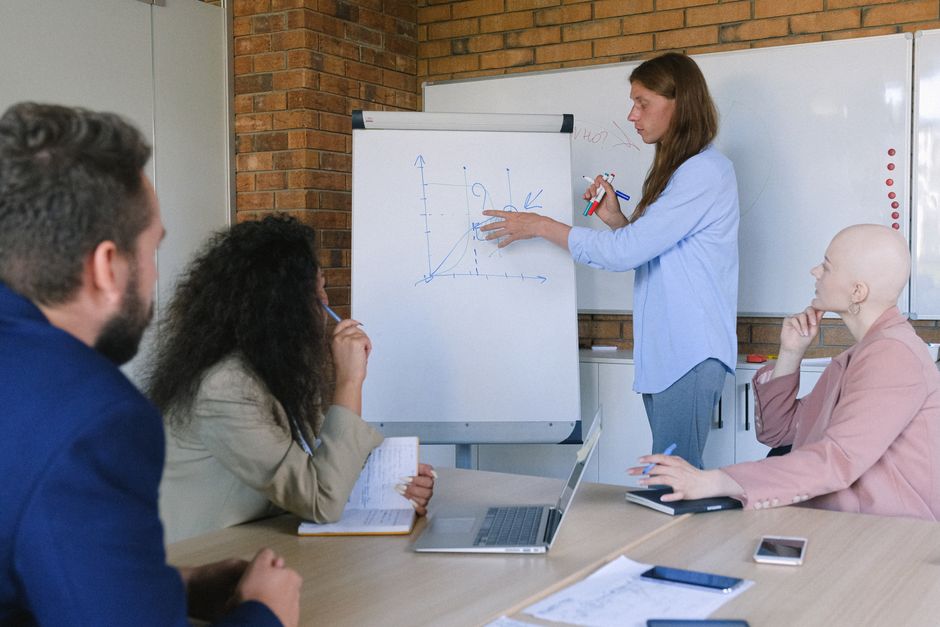 Düşünen bir grup iş arkadaşı, modern ofiste tahtada grafik çizen bir sunumu dinliyor.