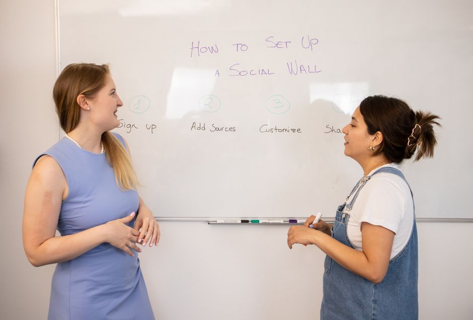 Two women discussing social media wall setup with tips on a whiteboard.
