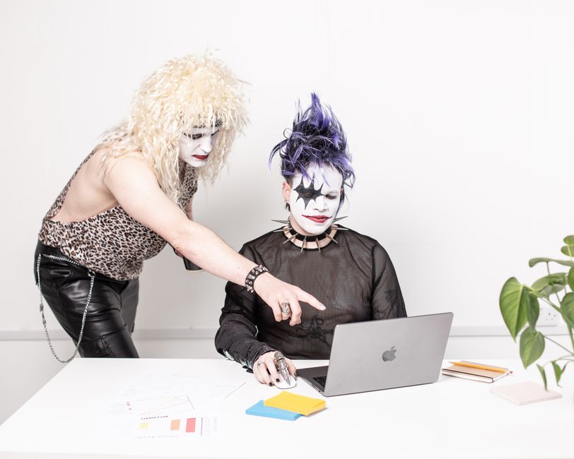 Two individuals with glam rock makeup using a laptop, blending creativity with technology.